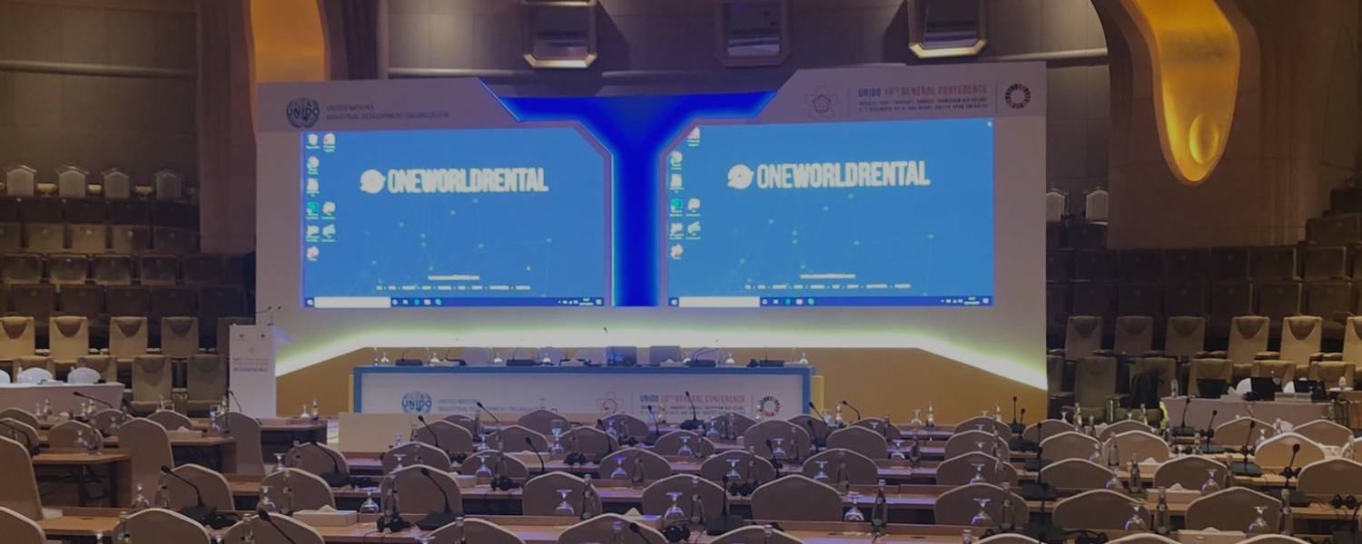 Large conference hall setup featuring rows of seating and a wide stage with dual screens displaying One World Rental branding.